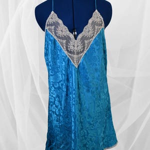May include: A turquoise satin slip dress with a floral pattern. The dress features a V-neckline with a lace insert and thin spaghetti straps. The hemline is trimmed with a gray border. The dress is displayed on a blue mannequin.