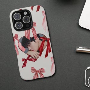 May include: A white phone case with a heart-shaped cutout displaying a person's image, adorned with red ribbon details. The case has a glossy finish and a camera aperture. The design incorporates pink watercolour bows and red and white striped bows.