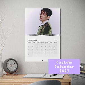 May include: A wall calendar for February 2027 featuring a portrait of a person with dark hair wearing a white collared shirt and a green velvet jacket. The calendar grid displays the days of the month clearly with black text on a white background.