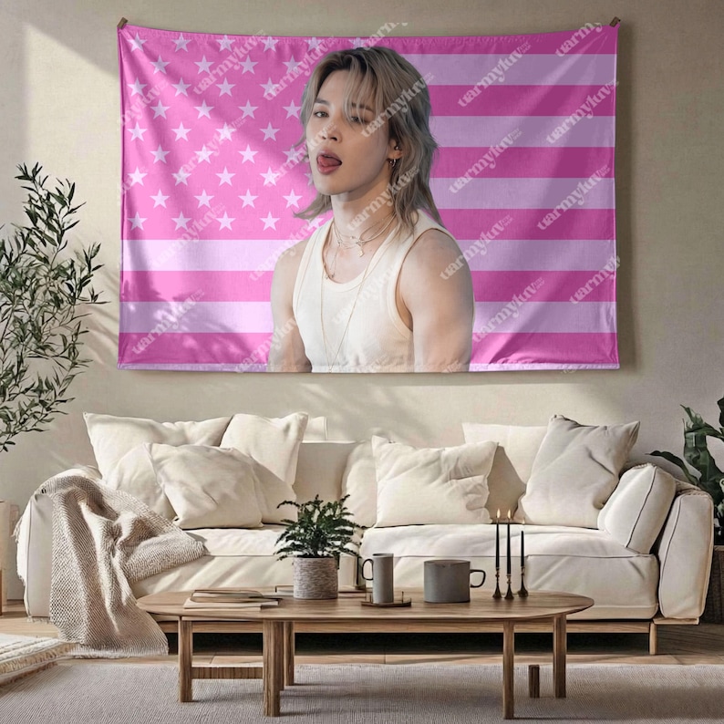 May include: A pink tapestry featuring a portrait of a person with light hair and a white tank top, set against a pink American flag design. The tapestry is hanging above a white sofa and wooden coffee table.