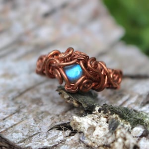 Op de afbeelding: Een ring van gewikkeld koperdraad met een kleine, iriserende blauwe edelsteen in het midden. Het metaalwerk bestaat uit ingewikkelde, kronkelende lussen en spiralen die de steen stevig op zijn plaats houden en een textuurrijke band vormen.