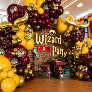 May include: A festive party scene with a brick wall backdrop, adorned with balloons in burgundy, gold, and yellow. The words "Wizard Party" are displayed, along with a Hogwarts Express sign, creating a magical atmosphere.