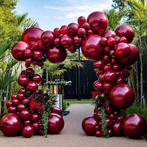 May include: A large, arched balloon installation composed of numerous glossy, deep burgundy balloons of varying sizes. The structure forms a decorative gateway, accented with small clusters of dark foliage and greenery at the base.