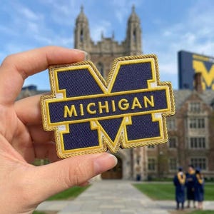 May include: Embroidered patch featuring the University of Michigan's block M logo. The patch is navy blue with gold trim and the word "MICHIGAN" in gold lettering. The patch is held up in front of a university building.