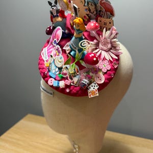 May include: A circular fuchsia fascinator hat adorned with various Alice in Wonderland themed miniatures including a figurine of Alice, a pocket watch, playing cards, a pink flamingo, mushrooms, a teapot, and small buttons on a fabric base.