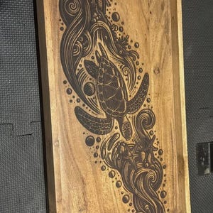 Puede incluir: Bandeja de madera con un diseño grabado con láser de una tortuga marina nadando entre olas y burbujas estilizadas. La bandeja es rectangular, con un borde elevado y un acabado de veta de madera natural.