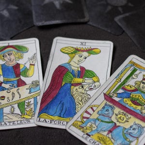 Puede incluir: Tres cartas de tarot con ilustraciones coloridas y títulos en francés. Las cartas representan figuras con trajes elaborados. Una carta muestra a una persona en una mesa, otra a una mujer con un león y la tercera a una persona en un carro.