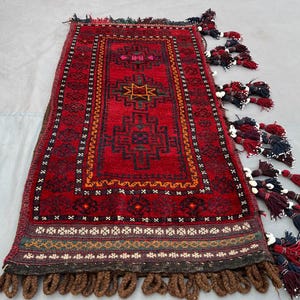 May include: A rectangular, hand-woven rug with a deep red field and intricate geometric patterns in black, orange, and yellow. The rug features a central medallion and is trimmed with tassels in shades of red, brown, and blue.