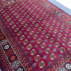 May include: A large, rectangular rug with a deep red background and intricate geometric patterns. The design features repeating diamond and floral motifs in shades of orange, white, and dark green, framed by a detailed border.
