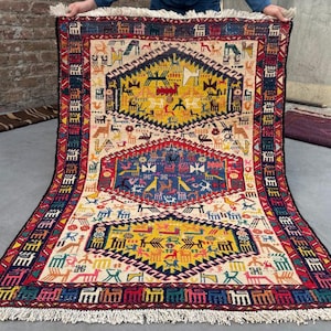 Puede incluir: Una alfombra rectangular tejida a mano con un campo de color crema y diseños geométricos en amarillo, azul, rojo y negro. La alfombra presenta un medallón central y bordes con patrones repetidos y flecos.