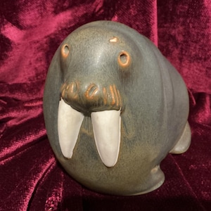 May include: A ceramic walrus figurine with a matte, dark gray-blue finish. The sculpture features two prominent, smooth white tusks, textured brown whiskers, and small, circular eyes. There is a small, light-colored chip on the top of the head.