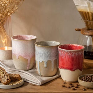 May include: Three ceramic tumblers with a reactive glaze finish. The left tumbler features a soft pink drip pattern over a beige base. The center tumbler has a white drip design on a tan body. The right tumbler displays a vibrant red drip glaze.