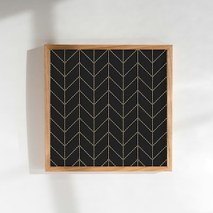 Op de afbeelding: Vierkant ingelijst kunstwerk met een zwarte achtergrond en een herhalend gouden chevronpatroon. Het kunstwerk zit in een natuurlijke houten lijst en biedt een moderne, minimalistische uitstraling. Geschikt voor diverse interieurstijlen.