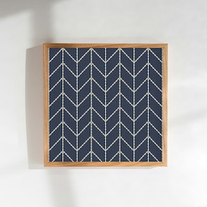 Op de afbeelding: Vierkant ingelijst kunstwerk met een marineblauwe achtergrond en een herhalend visgraatpatroon in gebroken wit. Het kunstwerk is ingekaderd in een natuurlijke houten lijst. Geschikt voor interieurdecoratie.