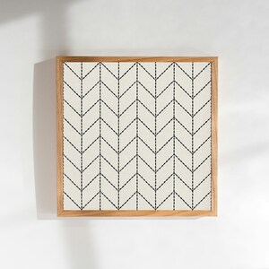 Op de afbeelding: Vierkant ingelijst kunstwerk met een crèmekleurige achtergrond en een herhalend zwart chevronpatroon. Het kunstwerk is ingelijst in een natuurlijke houten lijst, wat een minimalistische en moderne uitstraling creëert. Geschikt voor interieurdecoratie.