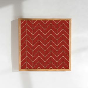 Op de afbeelding: Een ingelijst kunstwerk met een rode stof met een herhalend visgraatpatroon. Het patroon is gemaakt met goudkleurige stiksels. De lijst is gemaakt van lichtgekleurd hout en het kunstwerk wordt tegen een witte achtergrond gepresenteerd.