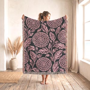 May include: A rectangular woven throw blanket featuring a dark charcoal background with a repeating pattern of large, detailed dusty pink dahlia flowers and leafy stems. The blanket has a decorative fringed edge along the top and bottom sides.