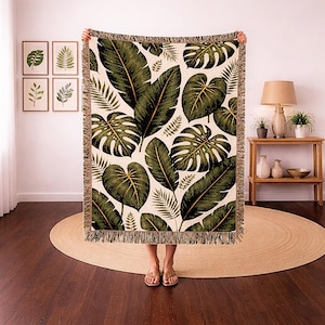 May include: A rectangular woven throw blanket featuring a repeating pattern of large green tropical leaves on a cream background. The blanket has a fringed edge and is held up by a person. The blanket is approximately 60 inches wide and 80 inches long.