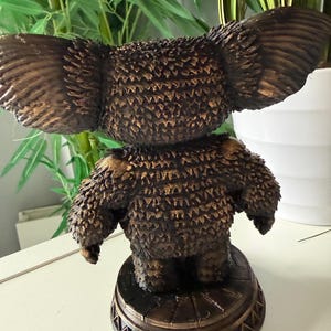 May include: A bronze-colored koala bear figurine with large ears and textured fur. The koala is standing on a decorative, round base with a geometric pattern. The figurine is set against a backdrop of green foliage and a white pot.