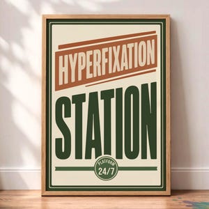 May include: A rectangular wall art print in a light wood frame featuring a cream background with a dark green border. The text HYPERFIXATION is in a rust-colored banner, and the word STATION is in large dark green letters with a circular 24/7 logo.