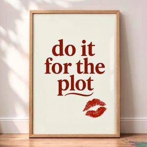 May include: A framed wall art print featuring the text "do it for the plot" in a bold, dark red serif font centred on a cream-coloured background. Below the text, there is a detailed red lipstick kiss mark graphic in the bottom right corner.