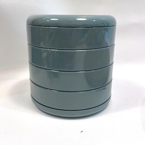 May include: A cylindrical storage container with a glossy, muted blue-green finish. The object features a stackable design composed of four distinct, rounded horizontal segments that fit together to form a single, uniform, smooth-surfaced unit.