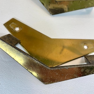 May include: Close-up of several vintage brass furniture legs. The legs have a distinctive angular design with a slightly aged, tarnished appearance. Each leg features a mounting hole for easy attachment.