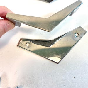 May include: A set of silver-toned, V-shaped furniture legs. One leg is held by a hand. The legs have a shiny, metallic finish and are designed with a modern, angular aesthetic. The legs have a length of approximately 5 inches.