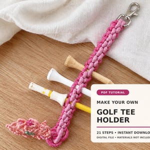 Op de afbeelding: Een roze macramé golftee-houder met een zilverkleurige metalen karabijnhaak aan de bovenkant. De houder is geweven met geknoopt koord en bevat drie golftees in de kleuren wit, geel en naturel hout. Een wit label toont de tekst PDF TUTORIAL MAKE YOUR OWN GOLF TEE HOLDER 21 STEPS INSTANT DOWNLOAD DIGITAL FILE MATERIALS NOT INCLUDED.