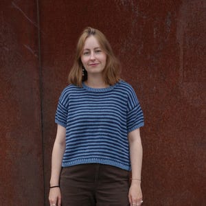 Könnte beinhalten: Ein kurzärmeliger Strickpullover mit horizontalen Streifen in abwechselnd hellblau und dunkelmarineblau, kombiniert mit einer einfarbigen braunen Hose und schwarzen Sportschuhen mit silbernen Akzenten.