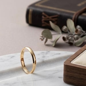 May include: A gold wedding band sits on a marble surface, with a blurred background featuring a closed book and a wooden ring box. The ring is simple and elegant, reflecting light.