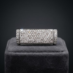 May include: A rectangular silver ring with a surface covered in small, sparkling gemstones. The ring is displayed on a gray velvet surface against a dark background, highlighting its brilliance and design.