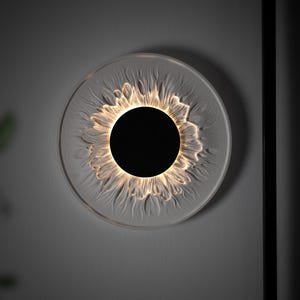 May include: A circular wall light fixture featuring a solid black center disc surrounded by a translucent, textured glass or acrylic ring that mimics the appearance of flower petals, illuminated from within to create a soft, warm glow.