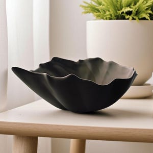 May include: A black decorative bowl with a wavy rim, resting on a light-colored wooden table. The bowl has a unique, organic shape and a matte finish. A white pot with a green plant is in the background.
