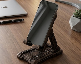 Może przedstawiać: Brązowy stojak na smartfona o futurystycznym designie, trzymający czarny smartfon. Inne przedmioty na drewnianym biurku to laptop, długopis, notatnik i słuchawki. Stojak ma około 10 cm długości i 8 cm szerokości.