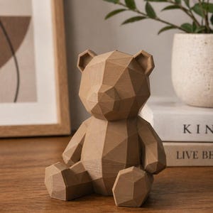 May include: A geometric, brown teddy bear figurine with a faceted design. The bear is sitting on a wooden surface, with a framed artwork and a potted plant in the background. The bear is approximately 6 inches tall.
