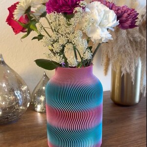 May include: A colorful vase with a wavy design, filled with a vibrant bouquet of flowers. The flowers include red, white, and purple blooms, with delicate white filler flowers. The vase is striped with pink, teal, and blue.