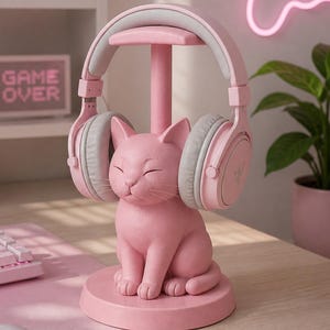 May include: A pink cat-shaped headphone stand featuring a smooth, matte finish and a circular base. The stand supports a pair of light pink and grey over-ear headphones with a sleek design, resting securely on the cat's head and ears.