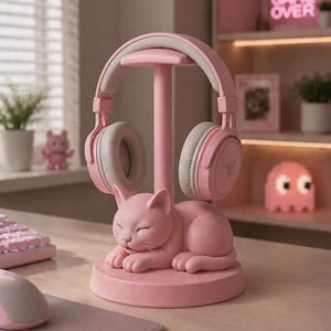 May include: A pink cat-shaped headphone stand featuring a sleeping feline figure at the base with a central vertical post. A pair of light pink over-ear headphones with grey ear cushions rests on the top of the stand, displaying a small logo.