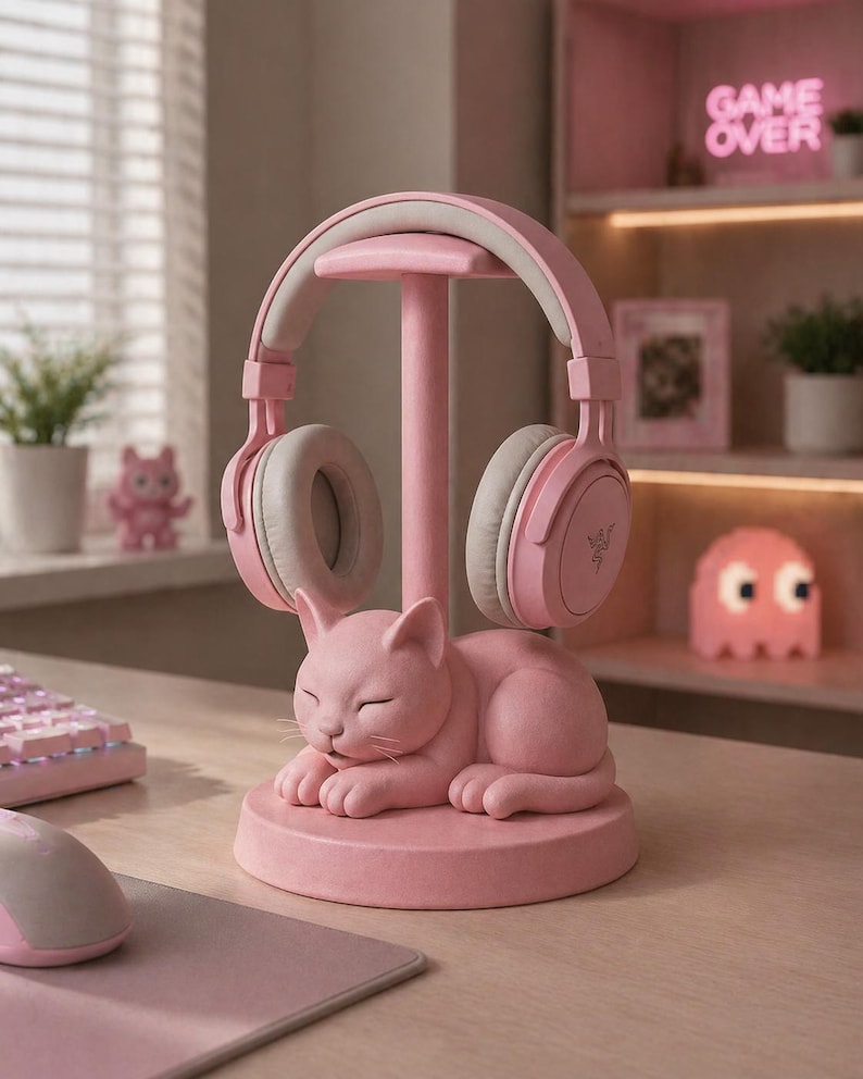 May include: A pink cat-shaped headphone stand featuring a sleeping feline figure at the base with a central vertical post. A pair of light pink over-ear headphones with grey ear cushions rests on the top of the stand, displaying a small logo.