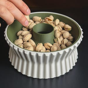May include: A white and green pistachio bowl with a fluted design, filled with pistachios. The bowl has a central compartment and is set against a dark background. A hand is reaching into the bowl to grab a pistachio.