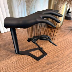 May include: A black metal sculpture of a hand holding a figure suspended by strings. The figure is in a reclining pose. The sculpture is on a wooden surface.