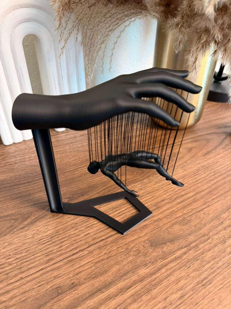 May include: A black metal sculpture of a hand holding a figure suspended by strings. The figure is in a reclining pose. The sculpture is on a wooden surface.