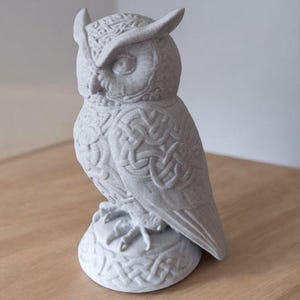 May include: A detailed, gray owl figurine with intricate Celtic knot patterns. The owl has large eyes, pointed ear tufts, and is perched on a round base. The statue is made of a textured material and stands on a wooden surface.