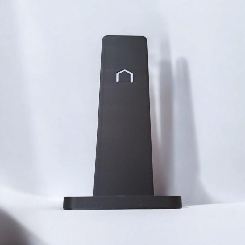 Puede incluir: Un soporte negro minimalista con base rectangular y cuerpo alto y c&oacute;nico. Un logotipo blanco en forma de casa est&aacute; centrado en la parte superior. El soporte est&aacute; dise&ntilde;ado para sostener un tel&eacute;fono o tableta.