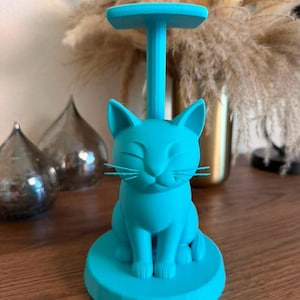 May include: A bright turquoise cat-shaped stand featuring a seated feline figure on a circular base with a vertical post extending upward to a flat top platform. The cat has closed eyes, pointed ears, and thin whiskers, all in a solid matte finish.
