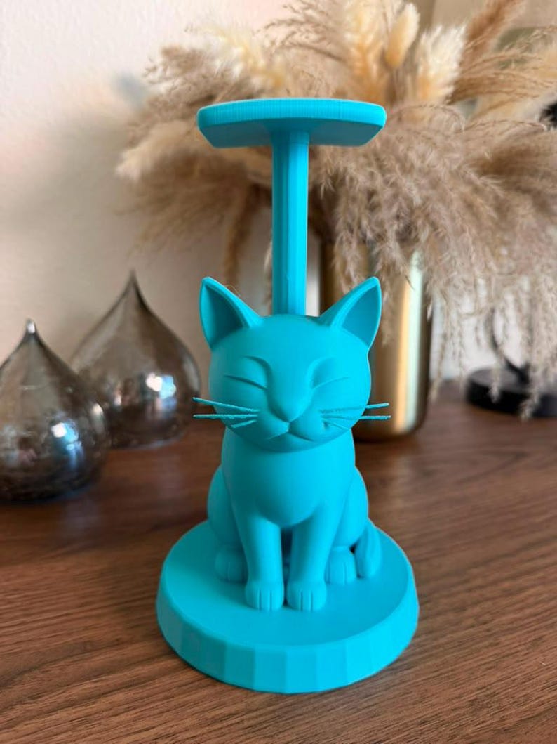 May include: A bright turquoise cat-shaped stand featuring a seated feline figure on a circular base with a vertical post extending upward to a flat top platform. The cat has closed eyes, pointed ears, and thin whiskers, all in a solid matte finish.
