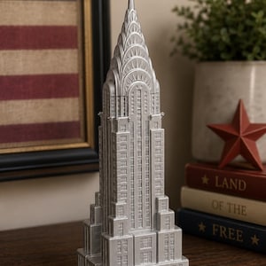 May include: A silver-colored model of the Chrysler Building featuring a tiered, stepped-back architectural design with a pointed spire, intricate window detailing, and a metallic finish, standing on a square base as a decorative sculpture.