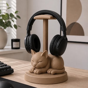 May include: A tan-colored headphone stand featuring a sculpted, sleeping cat at the base. The stand has a central vertical post with a horizontal top bar designed to hold a pair of black over-ear headphones securely in place on the cat's back.
