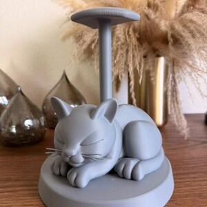 May include: A light gray paper towel holder featuring a sculpted sleeping cat on a circular base. The cat has closed eyes and visible whiskers, with a vertical pole extending from its back to support a horizontal top disc for holding rolls.